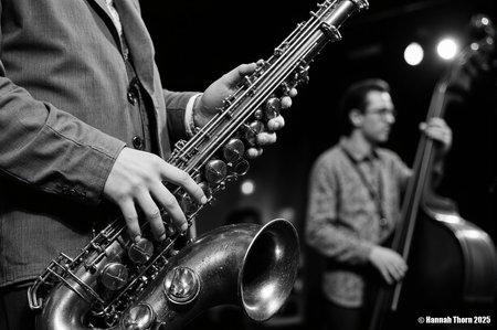 The image is a black and white close-up shot of a saxophonist playing a tenor saxophone.  The focus is sharply on the saxophone and the musician's hands, which are positioned on the instrument's keys and valves.  The saxophone itself appears aged, with a slightly worn look to its metallic surface. The details of the instrument, such as its keys and bell, are clearly visible.  The saxophonist is wearing a somewhat loose-fitting jacket and a visible portion of a collared shirt underneath. The background is significantly out of focus, but shows a blurry figure, seemingly another musician playing an upright bass, suggesting a jazz or blues performance setting.  The background lighting is dim, with some blurred light sources visible, contributing to the moody atmosphere.  The style of the photograph is distinctly **black and white photojournalism** or **concert photography**.  The high contrast, shallow depth of field (emphasizing the saxophone while blurring the background), and the candid feel (as if capturing a moment during a live performance) are all typical of these styles. The monochrome palette enhances the dramatic effect and focuses attention on the texture and form of the saxophone and the musicianâs interaction with it. The overall mood is intimate and intense, conveying the emotion and energy of a live music performance. <lora:_Flux\Vintage_Jazz-Club.safetensors:1.0:1.0>
