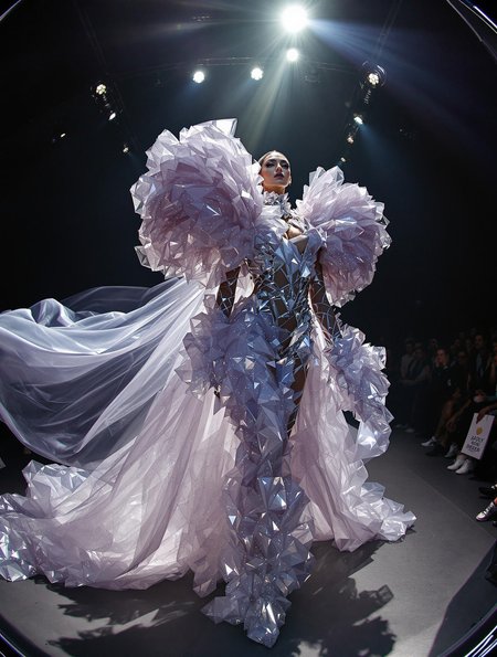 A surreal runway scene shot from a fisheye lens, the model wearing a crystalline couture outfit, oversized shoulders and flowing fabric trailing behind, deep shadows and studio lights, experimental fashion photo