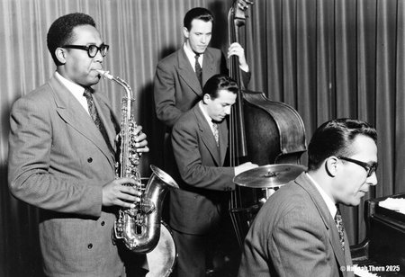 The picture is a black and white photograph of the David Brubeck jazz quartet performing.  The style is reminiscent of mid-20th-century jazz photography, likely taken sometime between the 1950s and 1960s. The monochrome palette, the slightly grainy texture, and the focus on the musicians in a seemingly informal performance setting all contribute to this classic jazz aesthetic. The image has a candid feel, not posed or overly staged, suggesting it may be a captured moment from a live performance or rehearsal. The clothing of the musicians (suits and ties) also supports the era's style. The photograph shows four musicians: * A saxophonist (likely alto saxophone) in the foreground on the left, prominently featured and playing his instrument.  He wears glasses and a suit.  * A pianist, also in the foreground to the right, is seated at a piano and appears to be playing. He also wears glasses and a suit.  * A double bassist stands behind the pianist, playing his instrument.  * A drummer is partially visible behind the pianist, seated at his drum kit.  Only the top of his head and shoulders are seen.   The background is somewhat blurry, but shows a curtain or drapery, common in performance venues. The overall composition focuses on the musicians and their instruments, making it a strong visual representation of a jazz performance.  The lighting is primarily focused on the musicians, creating a dramatic effect and drawing attention to their expressions and the music-making. <lora:_Flux\Vintage_Jazz-Club.safetensors:1.0:1.0>
