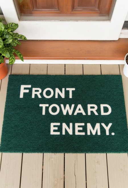 Photograph of a colorful welcome mat placed in front of a front door. The mat features a simple dark green design with the phrase "Front toward enemy" in bold, white, all caps letters.