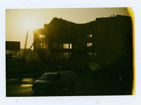 p0l2rd, Silhouette of a partially demolished building in the foreground, illuminated by the golden glow of the setting sun. The structure is surrounded by a haze of dust, with beams of sunlight piercing through. Visible cranes and machinery contribute to the industrial scene. The street below shows a billboard, an ambulance, and a few scattered vehicles. The sky is clear with faint contrails, underexposed. prominent film grain, high contrast lighting casting dramatic shadows, grainy film-like texture, polaroid-style format with white border, distinctive burnt edge, photograph appears aged or partially developed, with the right side fading into white