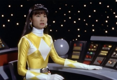 bridge of spaceship Star Trek Enterprise 1967, Trini wearing yellow power ranger catsuit, alone, long straight hair tucked behind shoulders, bangs, helmetless, uniform, white turtleneck collar, white gloves belt, holstered weapon, large chunky steel hoop earrings, worried facial expression, being transformed into a small porous cuboctahedron, sitting slightly-side-on, both arms very-outstretched, legs together, looking off-camera, in front of computer console on bridge of spaceship Star Trek Enterprise, Star Trek television series 1967, hyperdetailed photography