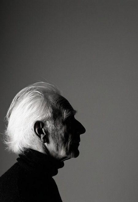 A dramatic black and white portrait photograph of an elderly man in profile, captured against a plain gradient background. The subject wears a black turtleneck sweater and stands with dignified posture, his white hair and pronounced profile creating a striking silhouette. The image employs side lighting and medium framing to emphasize the subject's distinguished features and create a powerful character study. Black and white portrait photography with sharp side lighting and high contrast medium shot composition.