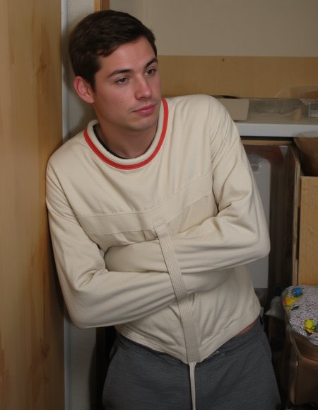 Medium shot of a young man in a small college dorm room. The man is handsome with defined jawlines, with short brown hair, and light freckles. He is wearing a white sjposey and grey sweatpants. He is kneeling, looking at the camera. His facial expression is neutral, almost serene, his gaze focused on a point beyond the camera. 
 <lora:straitjacketflux-posey:1.1>  <lora:amateurphoto-6version:0.8>