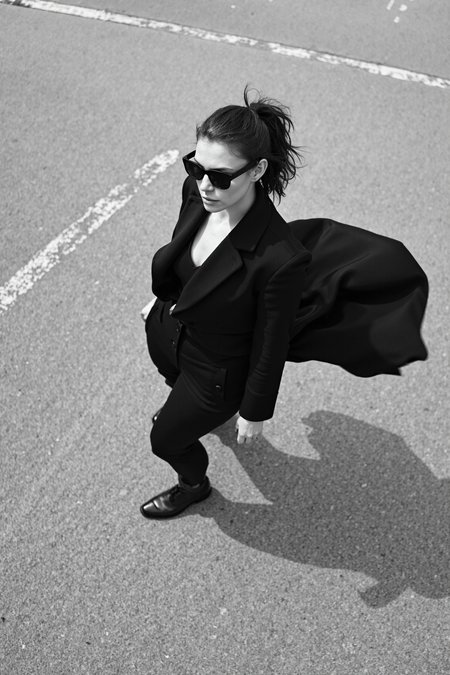sony_a7III professional style photo, Black and white photo of a stylish woman, early 20s, medium build, ponytail haircut, wearing angular black sunglasses and a flowing dark coat, mid-twirl creating dynamic motion in the fabric, standing on a textured concrete surface with faint painted lines, strong shadows and light interplay casting dramatic patterns, high contrast lighting accentuating the minimalist and cinematic mood, overhead perspective enhancing depth and movement in the composition