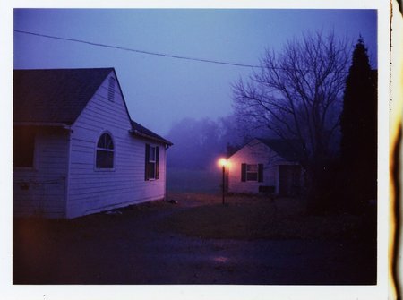 p0l2rd, Residential neighborhood at dusk, foggy evening setting, two houses visible, left house larger, white exterior, arched window, dark roof, right house smaller, grey exterior, black shutters, streetlamp glowing orange on the left side, bare trees silhouetted in the background, dark mailbox, empty driveway, dim lighting conditions, purple-blue haze dominates, outdoor urban environment, low visibility, street and grass barely discernible, overcast sky.  prominent film grain, high contrast lighting casting dramatic shadows, grainy film-like texture,  polaroid-style format with white border, distinctive burnt edge, photograph appears aged or partially developed, with the right side fading into white