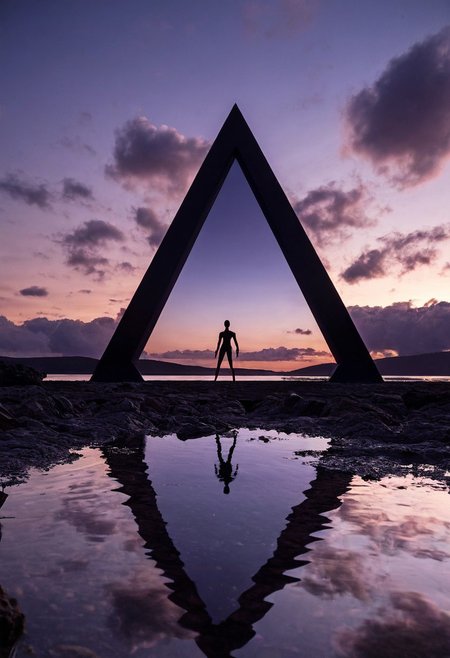 A colossal triangular architectural structure with intricate geometric cutout patterns emerges from a still body of water. The structure is constructed from dark materials and rises against a twilight sky filled with scattered clouds. A single human figure in complete silhouette stands motionless on a rocky shoreline in the foreground. The entire structure is reflected in the mirror-like water surface, creating a perfect mirror image. The sky features gradients of deep blue and purple hues, while the water surface reflects these colors, creating a symmetrical composition. The monumental scale of the triangular form dominates the frame, with its angular geometry and precise cutout patterns clearly visible against the evening sky. Low-angle architectural photography with natural backlighting and deep depth of field shows sharp detail from foreground to background.