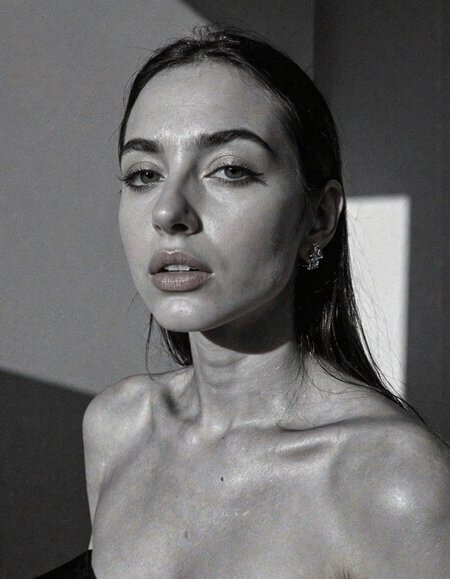 A striking monochrome portrait of a high-fashion model captured in a classic, Vogue-inspired photoshoot. The image focuses on her upper body, showcasing sharp bone structure and flawless skin illuminated by dramatic, high-contrast lighting. Shadows and highlights play artfully across her face, accentuating her high cheekbones, defined jawline, and elegantly arched eyebrows. Her gaze is powerful and direct, exuding confidence and sophistication, with slightly parted lips that add a touch of allure.

Her hair is styled sleekly back, drawing attention to her expressive features and creating a sculpted silhouette against a soft, shadowed background. She wears minimal accessories—perhaps a single, elegant earring catching the light—adding sophistication without distraction. The monochrome palette enhances the texture and contrast, giving her skin a smooth, almost ethereal glow, while the deep shadows add depth and mystery. The overall composition feels timeless and refined, embodying classic beauty and editorial poise with a touch of modern edge.. backinblack, grayscale, monochrome