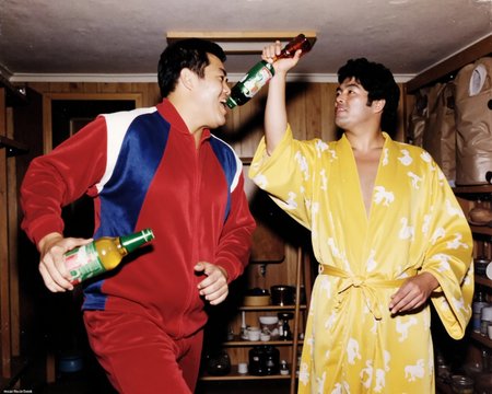 old color photo, a puroresu locker room scene where (25 year old man Jumbo Tsuruta in a red and blue velour track suit) is hurling beer bottles at (extremely tall dark-haired lanky with toothpick limbs wrestler Giant Baba in a robe) hitting him in the face getting beer all over his nice canary yellow Japanese silk robe with all over duck pattern print. In motion., analog, faded