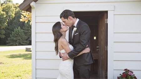 Candid photography of a couple embracing and kissing in front of a charming country chapel. The bride is wearing a stunning white wedding dress with intricate lace details, and the groom is dressed in a classic black tuxedo. They are standing in front of the small, wooden chapel on a sunny day. The bride's arms are wrapped around the groom, and his hands are gently placed on her waist. The soft sunlight illuminates the scene, creating a romantic and joyful atmosphere. The chapel and nature surrounding them add to the peaceful and loving moment. <lora:Sherry-00:1><lora:SelestaFlux1:1>
