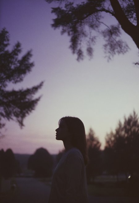 Film still, silhouette of a Girl Standing (out of focus) surrounded by trees, Twilight purple sky behind her,, Nikon F3, Nikon AF-NIKKOR 35mm f/2D Lens, kodak vision 3 500t Film, ISO 400, f/2.8, 35mm Film, Film Grain, film halation, Film look, Cinematic, epic cinematic color style