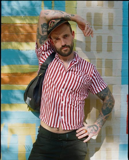 A man with a beard and tattoos standing in front of a colorful striped wall, posing confidently with one hand on his hip and the other resting on his thigh. he is wearing a red and white striped button-up shirt, black pants, and a black bag slung over his shoulder. his body is slim, and he has a beard. he has several tattoos on his arms, including one on his left arm and another on his right arm. he also has a cap on his head and a ring on his finger. the man is looking directly at the camera with a serious expression. the background is a vibrant outdoor setting with a striped wall in shades of blue, green, and yellow.
ðï¸ [Inpainted with Loras]: flux1-depth-dev-lora
ð¯<lora:flux\loratools\flux1-depth-dev-lora.safetensors:0.75:0.75>
