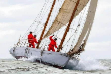 richardmacneil1 painting. A sailing boat with large sails navigates rough waters. Two people in red clothing adjust the sails and rigging. Waves crash against the boat. The sky is overcast. <lora:richardmacneil1_cap_d8a4e11:1.0><lora:853239920204979087:0>