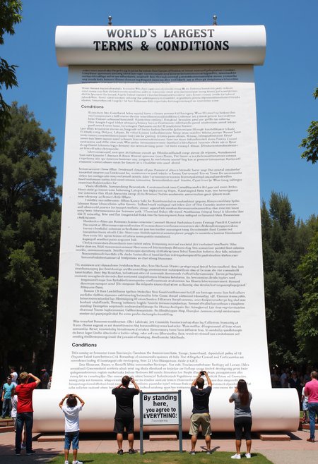 Photo depicting a skyscraper-sized paper scroll of dense legal jargon in a park that is titled, "World’s Largest Terms & Conditions". A sign at the bottom reads: "By standing here, you agree to EVERYTHING."  People stand around it taking pictures.