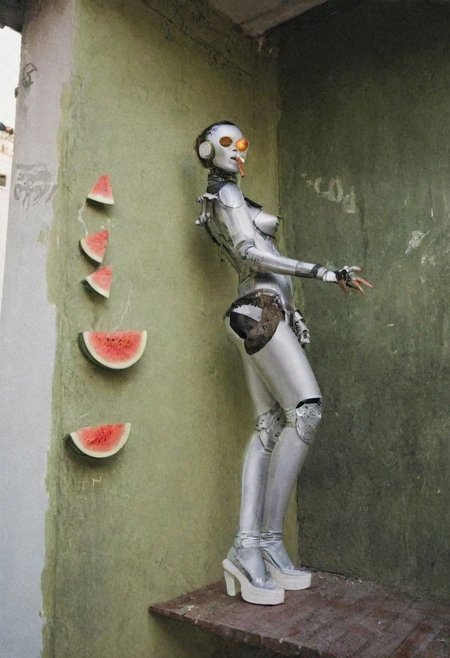 Editorial photograph, three gorgeous female brunette cyborg artists, dancing on the balcony of a typical messy, twenty-something's, So Paulo, Brazil apartment in the Liberdade barrio, a sassy but annoyed and slightly damaged gay robot stands in the corner smoking a cigarette, bad paintings of watermelon hang on wall, 1970s retrofuturism, dramatic moody cinematic lighting, Leica M3, film grain,