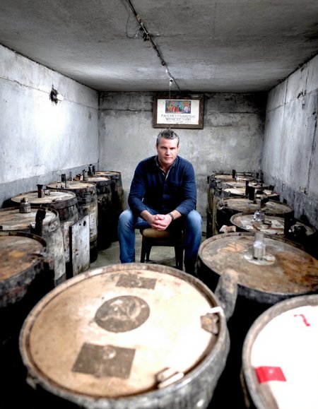 pete hegseth, a man, sitting alone in a concrete room surrounded by barrels of russian vodka marked with a hammer and sickle emblem. he is receiving signals from above.