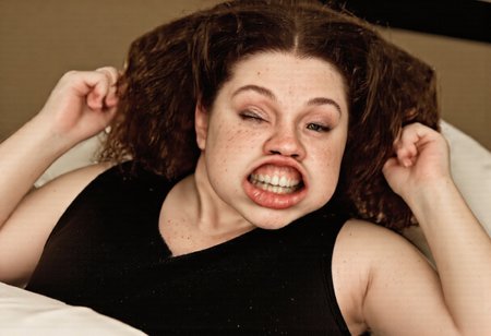 A medium shot photograph of a curvy woman with medium length curly hair standing laying down submissively in bed. hotel scenery. Night time, warm lighting. She has curly brown hair, lots of freckles on her nose and forehead, and brown eyes. She is wearing a black silk nightgown that exposes her cleavage. She has medium sized breasts that are very saggy. She has wide nostrils and a beautiful large smile. plump lips. w1nd8l0wn photo of a squinting woman's perfect teeth. windy. She is wearing clear lipgloss. big wide open mouth. stretchy flappy lips fully exposing gums and teeth. She is laying down in bed on a pillow. She has her arms above her head and to her sides in a submissive sexual pose. realistic. high-definition. She has a defined jawline and defined eyebrows. large bust. oval rectangular shaped head. si1k. sa1in. forehead freckles. nose freckles. Gina. shortstack.