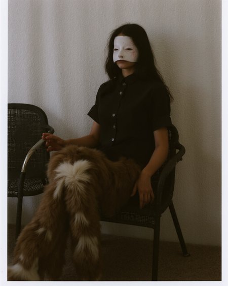 Kaos, The image is a photograph with a surreal, eerie atmosphere. The subject is a person, likely a young woman, with a face mask that obscures their features, giving them an unsettling, almost inhuman appearance. They have long, dark hair cascading down their shoulders. The person is dressed in a dark, short-sleeved, button-up shirt, which contrasts with the lighter, ethereal background. The individual is seated in a dark, wicker chair with a mesh backrest. Their legs, covered in fur, are positioned in a relaxed, slightly spread stance, with one foot resting on a chair. The fur appears to be a mix of brown and white, adding to the otherworldly feel of the image.