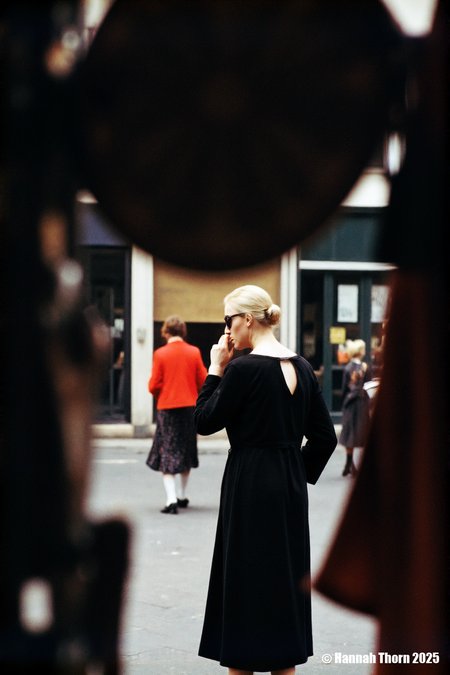The picture is a street photograph, likely a candid shot, capturing a moment in a public space.  A woman, seen from the back and slightly to the side, is the central figure.  She wears a simple, dark, possibly black, long-sleeved top or dress with a low v-back. Her blonde hair is pulled back, and she's wearing dark sunglasses. Her right arm is bent, and her hand is raised to her face, either touching her cheek or adjusting her glasses.  In the background, another woman, smaller and slightly out of focus, walks away from the camera. She's wearing a vibrant red jacket and a dark, patterned skirt that falls below the knee. Her legs are visible, and she appears to be wearing light-colored stockings and dark shoes.  The overall style of the photograph is reminiscent of street photography from the mid-20th century.  Several elements contribute to this impression:  * **The use of color:** The colors are somewhat muted, suggestive of older film stock.  The pop of red from the woman in the background contrasts with the darker tones of the central figure and the overall scene. * **The focus and depth of field:** The central woman is in sharper focus, while the background and the second woman are softer, creating a sense of depth and isolating the main subject. * **The framing and composition:** The photograph is cropped in a way that emphasizes the central woman while also including the other woman in the background as a secondary element of interest. The dark, undefined shapes in the foreground suggest the photographer may have been shooting from behind a partially obscured vantage point, like through a window or foliage, adding a layer of mystery and candidness. * **The grainy texture:** A noticeable grain or noise is present throughout the image, adding to the vintage feel and again hinting at the use of film. * **The moment captured:** The scene is everyday and unposed, characteristic of street photography's aim to capture fleeting moments of real life. The woman's gesture suggests a natural, unselfconscious action, rather than a posed portrait.  The photograph evokes a sense of mystery and intrigue, partly due to the obscured foreground, the candid nature of the shot, and the fact that the central woman's face is partially hidden by her hand and sunglasses.  The image invites the viewer to speculate about the context of the scene and the women's thoughts or actions. The style is observational and documentary, focusing on the interplay of light, shadow, and human presence in a public environment. It has a timeless quality that speaks to a particular era of street photography, perhaps the 1950s or 1960s. <lora:_Flux\Saul_Leiter_Streetphotography.safetensors:1.0:1.0>