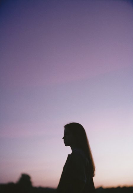 Film still, silhouette of a Girl Standing (out of focus), Twilight purple sky behind her,, Nikon F3, Nikon AF-NIKKOR 35mm f/2D Lens, kodak vision 3 500t Film, ISO 400, f/2.8, 35mm Film, Film Grain, film halation, Film look, Cinematic, epic cinematic color style