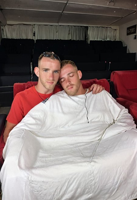 A man sleeping in a movie theater bench seat church pew with a blanket snuggled up next to another male. They are both covered up in a blanket, snuggled together, head on shoulder,