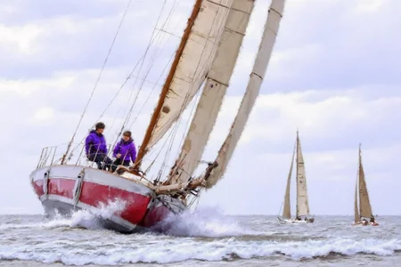 richardmacneil1 painting. A red and white sailboat navigates choppy waters with two people onboard wearing purple jackets. The sails are fully extended, and waves crash around the boat. In the background, additional sailboats are visible under a partly cloudy sky <lora:richardmacneil1_cap_d8a4e11:1.0><lora:853234130589091849:0>