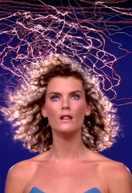 135mm shot on sony analog HDTV camera laserdisc style dorky australian brunette woman with thick curly hair in standing in large room with soft goo dripping off electric concrete with cables on floor like television studio wide shot surrounded by swirling colour and sexy smirking at camera with a little wink. background is 1990s japanese beauty scenes exploding televisions electricity in background in a surreal fashion technology moving into her head cables and wires low contrast super sharp imagery