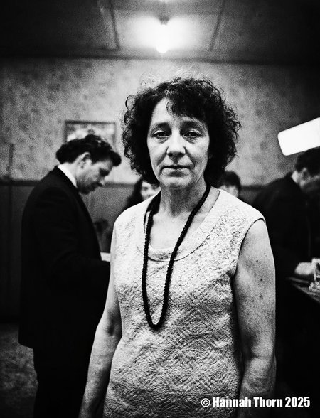 A black and white photograph from a portrait camera angle about a middle-aged woman with curly hair, wearing a knitted dress, standing in a dimly lit room with blurred figures in the background. the woman, who appears to be in her late 60s or early 70s, is standing in the middle of the image, with her upper body facing the viewer and looking directly at them. she has a serious expression and is wearing a sleeveless, lace-like dress with a black beaded necklace around her neck. her curly hair is styled in a way that frames her face and adds to her natural beauty. the background is out of focus, focusing attention on the woman and her outfit.<lora:_Flux\Anders_Petersen.safetensors:0.9:0.9>