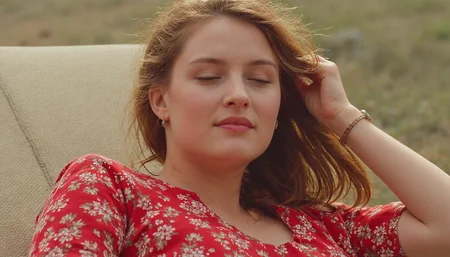 cinematic medium of a young woman reclining on a deck chair, wearing a red floral pattern summer dress, open hair blowing in the breeze, eyes closed, faint smile. Warm natural light. High resolution. 2020s movie <lora:dark_cinematic-000080:1>