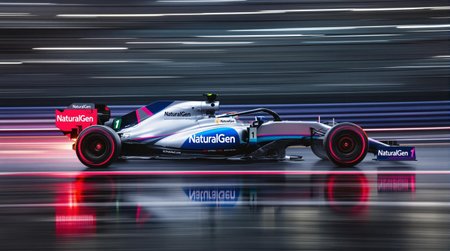 A sensational photograph photograph of an f1 car in motion, with glowing hot brakes.  The background and track and wheels are blurred in motion capturing the essence of speed.  The race car's brakes are glowing red hot, and are reflected on the wet track.  There is a bit of jitter.  The lighting is dramatic and creates high contrast, but the image is just a bit washed out due to age.  The formula one race car is painted in a delightful gray and blue and magenta scheme with the number 1 and the is sponsored by "NaturalGen".  The car is seen in profile, directly from the side.  The image is expertly composed and is suitable as an artistic work in minimalist technical photographic style