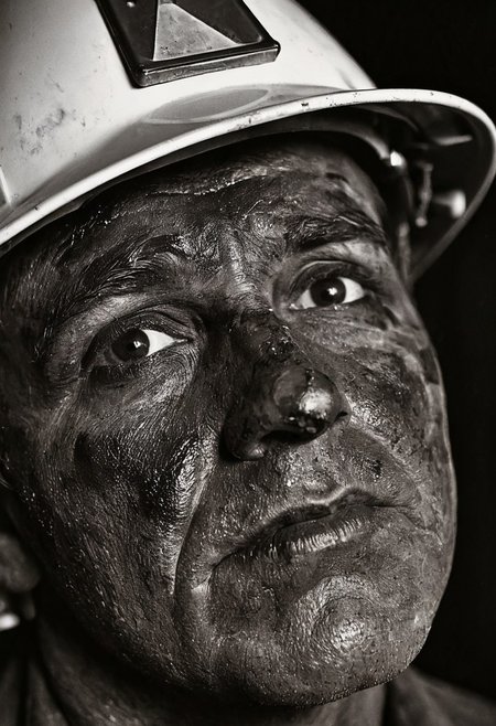 A close-up portrait photograph of a coal miner's face, focusing on his eyes beneath a coal-stained brow. His face is covered in black coal dust, creating stark contrast against the whites of his eyes. The miner wears a traditional hard hat with a headlamp, and his weathered skin shows traces of coal residue. The background is dark and out of focus, suggesting the underground mining environment. Black and white documentary photography with dramatic side lighting and sharp focus on facial features. 4k, intricate details, highly detailed, hyper-realistic, 8k RAW Editorial Photo. cinematic light, dramatic light, shallow depth of field, high budget, cinemascope film grain ISO 200 faded film, 35mm photo, grainy, vignette, vintage, Kodachrome,