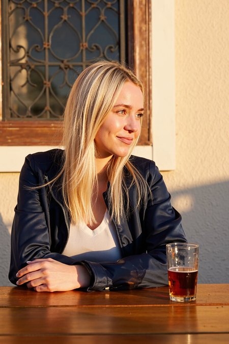 sony_a7III professional style photo, Female, early 20s, fair-skinned, slim build, long straight blonde hair neatly framing face, dark navy jacket partially zipped revealing light-colored inner layer, clear glass of amber beer with slight foam placed on a polished wooden surface, outdoor setting with rustic iron latticework window visible in the background, textured wall surface adding character to the environment, soft warm lighting casting a golden glow on the subject, subtle shadows creating depth, slight smile conveying relaxed expression, side gaze directed away from the camera, medium camera angle capturing upper body, warm ambient evening lighting enhancing natural tones, serene and inviting atmosphere, casual yet refined outdoor scene