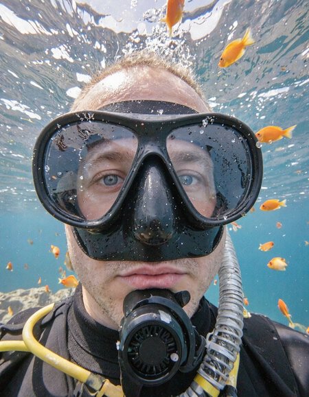 This is a vivid photograph depicting a close-up view of a diver's face underwater. The diver is wearing a full-face diving mask with a black frame and a clear visor. The visor is slightly fogged, adding a sense of realism to the scene. The diver's eyes are blue and wide open, reflecting the vibrant underwater environment. Water droplets cling to the visor, emphasizing the wet and aquatic atmosphere.

The background is a rich, deep blue, typical of an underwater setting. Scattered throughout the frame are bright orange and yellow tropical fish, adding a burst of color. The fish are small, with slender bodies and large eyes, swimming freely in the water. The clarity and sharpness of the image highlight the textures of the diver's mask and the fish's scales, giving a sense of depth and immersion.

The lighting is natural, likely from sunlight filtering through the water, creating a warm, inviting ambiance. The overall mood of the photograph is one of exploration and tranquility, capturing the thrill of diving in a tropical reef. The image is highly detailed, with a focus on the interplay between the human and the underwater world.