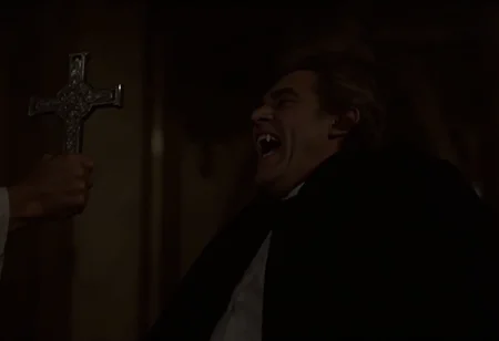 medium shot of a middle-aged aristocratic vampire man recoiling from a priest inside a gothic cathedral. The vampire is standing on the right side of the frame, hissing showing fangs, dramatically billowing his cape. The priest is standing on the left side of the frame, holding a sivler cross towards the vampire directly looking at him. soft warm light at night <lora:hammer-000073:1>