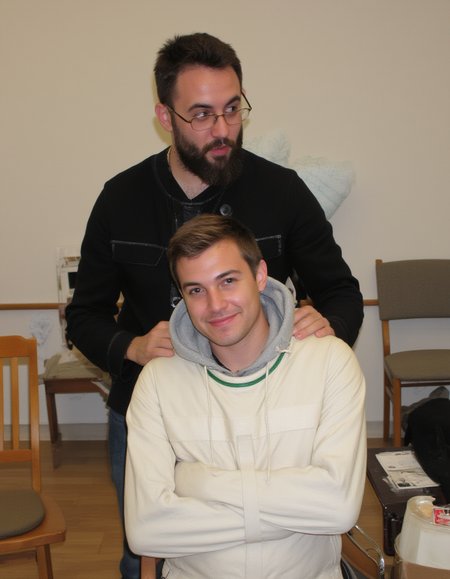 A photograph, likely in a classroom or meeting room. In the foreground, a stylish man with short hair and a beard, wearing a black open-button shirt and glasses, is standing and doing something behind second person. The second person, a handsome man with short hair, is wearing a white sjposey, with a grey hood behind his neck. He has a slight smile, suggesting a playful or joking mood. The room features a mix of wooden and upholstered chairs, adding to the casual atmosphere. <lora:straitjacketflux-posey:1.1> <lora:amateurphoto-v6-forcu:0.8>