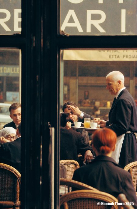 The image captures a scene within a Parisian café, viewed through a darkened vertical element, likely a door frame or window mullion. The focus is on a waiter, an older man with close-cropped white hair, dressed in a dark suit jacket and white apron, holding a tray laden with drinks and cups. He appears to be serving a group of patrons seated at wicker chairs.   The patrons are less distinct, partially obscured by the foreground element and the soft focus of the image.  A woman with a light-colored head covering and glasses is visible in the lower left corner, seemingly absorbed in something she holds in her hands.  Other figures are present, including a man with dark hair who is partially obscured by the frame, and a woman with reddish hair who appears to be raising her hand to her face.  The overall style of the image evokes a candid, street photography aesthetic. The muted color palette, dominated by browns, grays, and dark tones, contributes to a vintage or nostalgic feel. The soft focus and grainy texture, along with the slightly blurred background, suggest that the photo might have been taken with a slower shutter speed, perhaps capturing a fleeting moment in the café's life.    The composition, with the dark vertical bar in the foreground partially obscuring the scene, creates a sense of depth and voyeurism, as if the viewer is catching a glimpse of a private moment.  The blurred background, including what appears to be signage or lettering on the wall behind the waiter, adds to the impression of a bustling, lively atmosphere.  The lighting is diffuse, likely natural light filtering into the café, contributing to the overall ambiance of the scene.  The style could be described as realistic, capturing the everyday life of a Parisian café in a naturalistic manner. <lora:_Flux\Saul_Leiter_Streetphotography.safetensors:1.0:1.0>