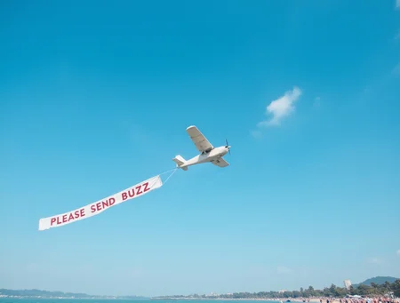 small white airplane towing a long banner that reads "PLEASE SEND BUZZ" across a vibrant, clear blue sky, flying over a crowded beach