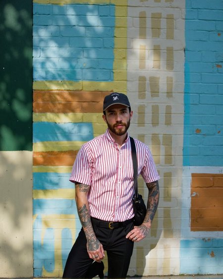A man with a beard and tattoos standing in front of a colorful striped wall, posing confidently with one hand on his hip and the other resting on his thigh. he is wearing a red and white striped button-up shirt, black pants, and a black bag slung over his shoulder. his body is slim, and he has a beard. he has several tattoos on his arms, including one on his left arm and another on his right arm. he also has a cap on his head and a ring on his finger. the man is looking directly at the camera with a serious expression. the background is a vibrant outdoor setting with a striped wall in shades of blue, green, and yellow.