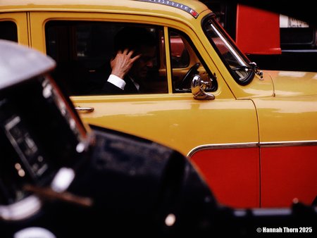 The image is a photograph, likely taken candidly on a city street. It features a vibrant yellow and red taxi, with the focus deliberately placed on a blurred foreground element, possibly another vehicle or object. Through the taxi's window, we partially see a passenger in a dark suit and white shirt, his hand raised to his face or head.  The rest of his features and the background inside the taxi are obscured by shadow and the relative brightness of the window's reflection. In the bottom left corner of the image, a blurred object, potentially a cigarette, is visible, adding a further layer of urban detail.  The style of the photograph is reminiscent of street photography, particularly work from the mid-20th century. It shares characteristics with the style of photographers like Saul Leiter, known for his evocative use of color, reflections, and layered compositions to capture fleeting moments of urban life. The blurred foreground element and the partially obscured subject create a sense of depth and mystery, drawing the viewer into the scene while simultaneously keeping them at a distance. The vibrant colors of the taxi contrast with the darker tones of the foreground and the shadowed interior, adding to the image's visual impact.  The photograph's focus is not on sharp detail but on capturing an atmosphere and a transient moment, suggesting a snapshot of life in motion.  The overall effect is one of both vibrancy and anonymity, capturing the bustle and anonymity of city life. <lora:_Flux\Saul_Leiter_Streetphotography.safetensors:1.0:1.0>