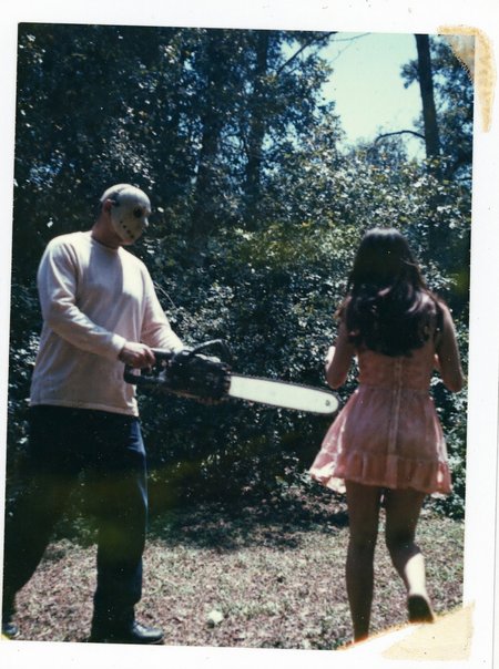 p0l2rd, Overexposed and color-distorted instant photo, man wielding a running chainsaw, facing towards a young woman running away, tall trees and dense foliage filling the background, man wearing a light-colored long-sleeve shirt, dark pants, dark shoes, and scary hockey mask. The woman with long dark hair, a light pink or coral dress, and dark shorts, the sunlight through the trees creates lens flares and hazy patches, outdoor forest setting, daytime, likely a staged or posed scene, damaged edges, colors shifted toward blue and green tones. prominent film grain, high contrast lighting casting dramatic shadows, grainy film-like texture