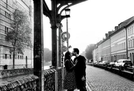monochrome, instagram photo, a loving couple is standing at a street lamp and kissing. this lantern is at the head of an old bridge. in the background you can see an old town with a castle and a church, which is foggy. it's autumn, foggy and it's at night