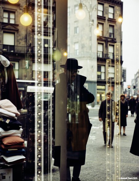 The picture is a photograph that appears to be taken through a shop window.    **Description:**  The main focus is a blurry, partially obscured figure of a person wearing a fedora hat, positioned centrally. This figure is reflected in a glass surface, which creates a double image effect.  The reflection is superimposed over a street scene visible through the shop window. The street scene shows piles of stacked merchandise, possibly clothing, with price tags or signs visible.  There's also a building in the background with architectural details suggestive of an older style of building. Several light bulbs are visible both inside the shop and reflected on the street outside. The overall atmosphere is one of urban life, possibly in a city during a cold season.  Other blurry figures of people can be seen moving in the street scene. The details of the street scene are not entirely clear due to the reflection and blur.   **Style:**  The style of the photograph is reminiscent of street photography, with a focus on capturing candid moments of urban life. The use of reflections and blurring adds an element of abstraction and mystery. It exhibits the characteristics of a slightly surreal, dreamlike quality due to the way the reflection interacts with the scene. The overall aesthetic is gritty, evocative of a certain era. The monochrome palette enhances this effect, emphasizing the contrasts in light and shadow. The photographer seems to be interested in exploring the intersection of the interior and exterior spaces through the medium of reflection. The style aligns with a modernist approach to photography, valuing observing and recording moments of contemporary life in an unconventional way.  There's a strong contrast between the sharpness of some elements (like the building) and the intentional blurriness of others (the people and the central figure), creating a layered depth and visual interest. <lora:_Flux\Saul_Leiter_Streetphotography.safetensors:1.0:1.0>