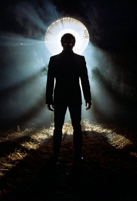 A shadowy silhouette with glowing eyes is standing in a dark room. The darkness of the room and the man's eyes create a striking contrast, drawing attention to the subject. The man is the main focus of the image, and his presence in the dark room adds an element of mystery and intrigue. 1970s horror film