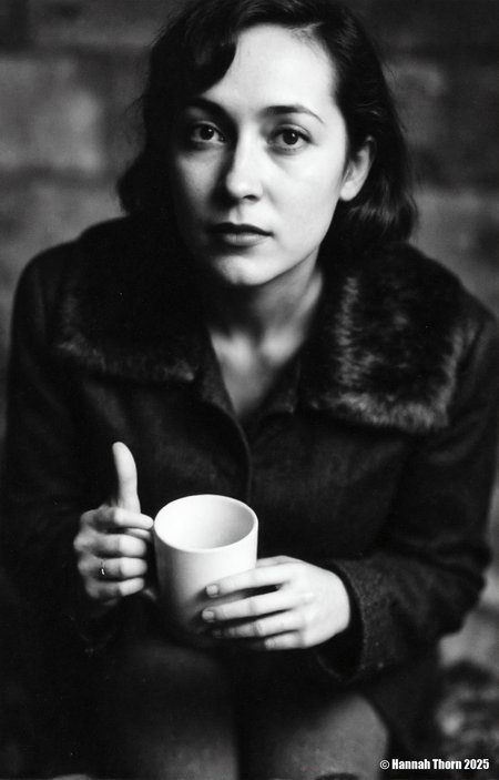 A black and white photo from a portrait camera angle about a woman holding a cup, looking directly at the viewer with a serious expression. the woman, who appears to be in her late 20s, has long, wavy hair and is wearing a black coat with a fur collar. she is sitting in the middle of the image, with her upper body visible, and her hands are holding a white mug. the background is blurred, with a dark, moody atmosphere, and the focus is on the woman's face and the mug she is holding. the image has a minimalist aesthetic, with no other elements present in the scene.<lora:_Flux\Saul_Leiter_BW.safetensors:1.0:1.0>