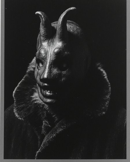 Kaos, The image is a black-and-white photograph  dressed in a fur-lined coat, which appears to be made of coarse, textured material, giving it a rugged, almost wild look. His face is obscured by a grotesque, grinning mask that is worn over his head, with elongated, curved ears that resemble a rabbit's ears. The mask is made of a material that reflects light, giving it a shiny, almost metallic texture. The mask's eyes are sunken and menacing, with a wide, menacing grin, adding to the eerie atmosphere.