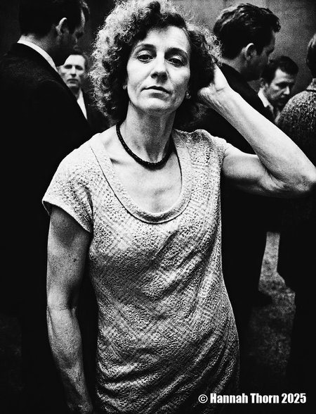 A black and white photograph from a portrait camera angle about a middle-aged woman with curly hair, wearing a knitted dress, standing in a dimly lit room with blurred figures in the background. the woman, who appears to be in her late 60s or early 70s, is standing in the middle of the image, with her upper body facing the viewer and looking directly at them. she has a serious expression and is wearing a sleeveless, lace-like dress with a black beaded necklace around her neck. her curly hair is styled in a way that frames her face and adds to her natural beauty. the background is out of focus, focusing attention on the woman and her outfit.<lora:_Flux\Anders_Petersen.safetensors:0.9:0.9>