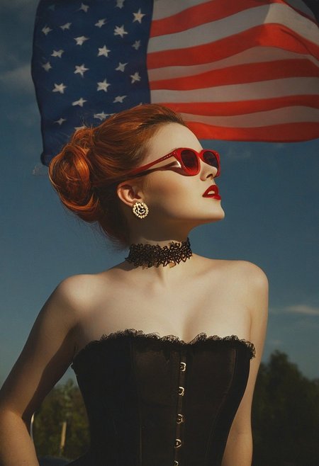 This photograph captures a striking woman in a dramatic, vintage-inspired pose against the backdrop of a sunset, with a car and an American flag in the background. The subject is a young woman with light skin and auburn hair styled in an updo adorned with a large, prominent red rose. She is wearing large, stylish, red-framed sunglasses, adding a touch of retro flair. Her attire includes a black, strapless, corset-style top that cinches at the waist, revealing her shoulders and hinting at a more modest bust size. 
Her jewelry consists of dangling earrings featuring small, ornate cherry charms. The choker she wears around her neck is black, adding to the edgy, gothic vibe. The lighting is dramatic, casting a warm, golden glow that enhances her porcelain complexion and adds depth to the image. She is caught in a side profile, her eyes closed, and mouth open, suggesting a moment of deep thought or a slight smile.
The background features a partially visible American flag to the left and a dark silhouette of trees, indicating an outdoor setting. The sky above is a mix of dark clouds and golden hues, suggesting the transition from day to night. The overall mood is one of vintage glamour with a touch of rebellious flair.
