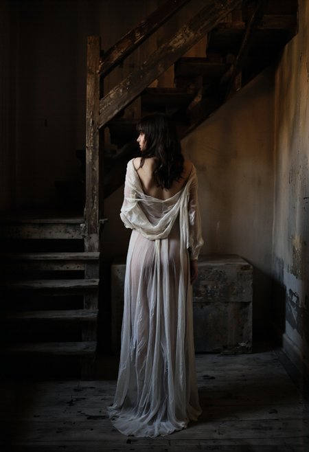 NyxBorne. A black and white photo shoot from behind about a woman standing in a dimly lit room, facing away from the camera, with her back towards the viewer. the woman, who appears to be in her mid-twenties, is standing in the middle of the frame with her full body visible, wearing a sheer white dress that is slipping off her shoulders. her long, dark hair cascades down her back, and she has a slender, toned physique. the room has a rustic, dilapidated appearance with wooden stairs in the background, and the lighting is soft and diffused, casting a warm glow on the woman's figure. the overall mood of the image is mysterious and alluring, with a focus on the subject's figure and the texture of her dress.