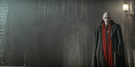 Horror-themed,  <lora:Fright Night 1985 Film Style:1>
Cinematic Wide Shot Film Footage captured by Panavision Cameras and Lenses, Vampire Creature Jerry vampire nosferatu in a long coat jacket and red scarf staring at camera in a dark basement room with dramatic dim light on, Fright Night 1985 Film Style, Eerie, unsettling, dark, spooky, suspenseful, grim, highly detailed