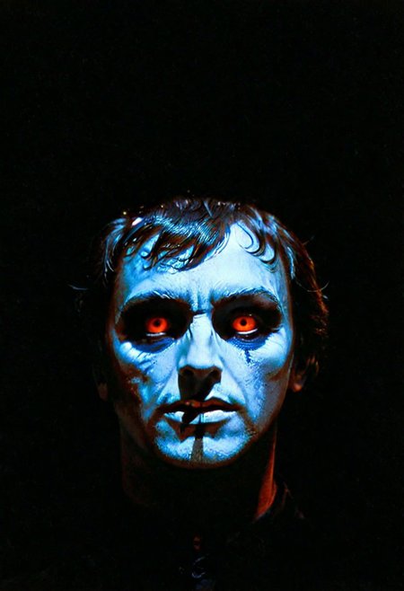 A shadowy silhouette with glowing eyes is standing in a dark room. The darkness of the room and the man's eyes create a striking contrast, drawing attention to the subject. The man is the main focus of the image, and his presence in the dark room adds an element of mystery and intrigue. 1970s horror film